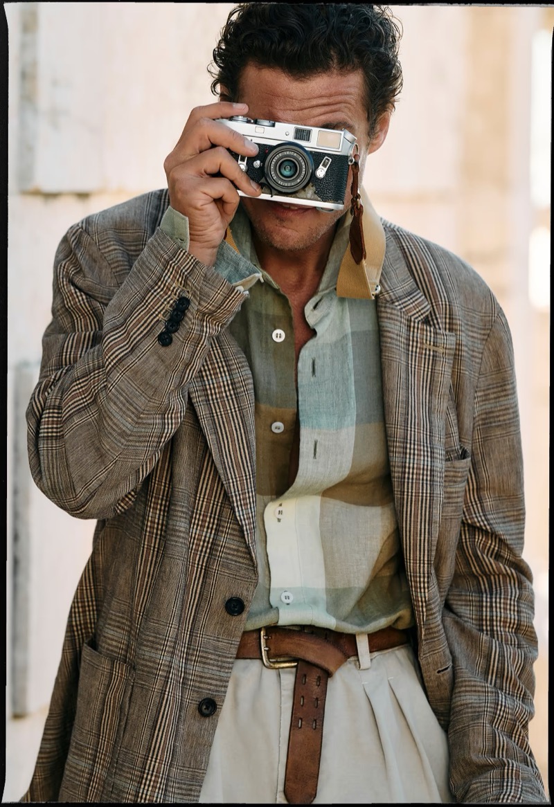 Jon Paul raises a silver Leica camera to his eye in a brown windowpane plaid linen blazer over a colorblock checked linen shirt with a thick leather belt and pleated cream trousers