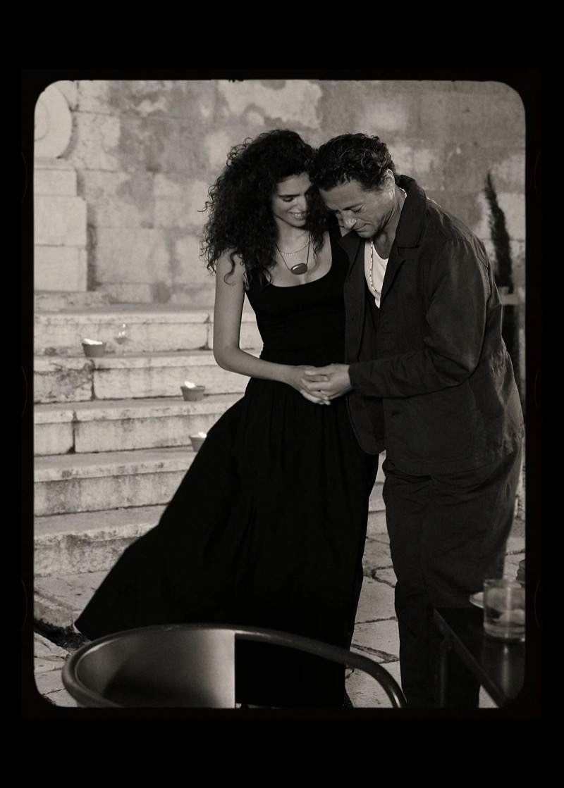 Black and white image of Jon Paul slow-dancing with a female model on candlelit stone steps in a dark linen jacket and dark trousers with a long beaded necklace
