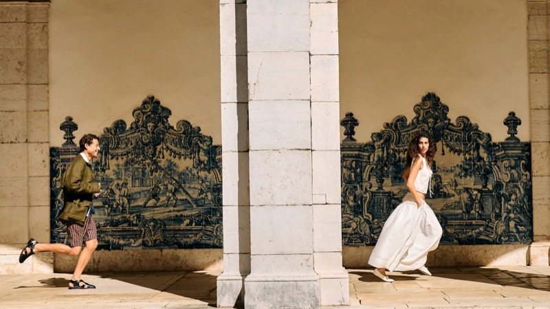 Jon Paul runs past blue and white azulejo tiles under a limestone arcade in an olive utility jacket with brown striped shorts and black loafers, mirrored across a column by a female model in a cream linen vest and skirt