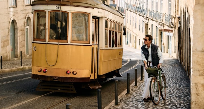 Jon Paul walks a green vintage bicycle along Lisbon cobblestones as a yellow tram passes behind him, wearing a slate gray quilted vest over a chambray shirt with white linen trousers and brown loafers
