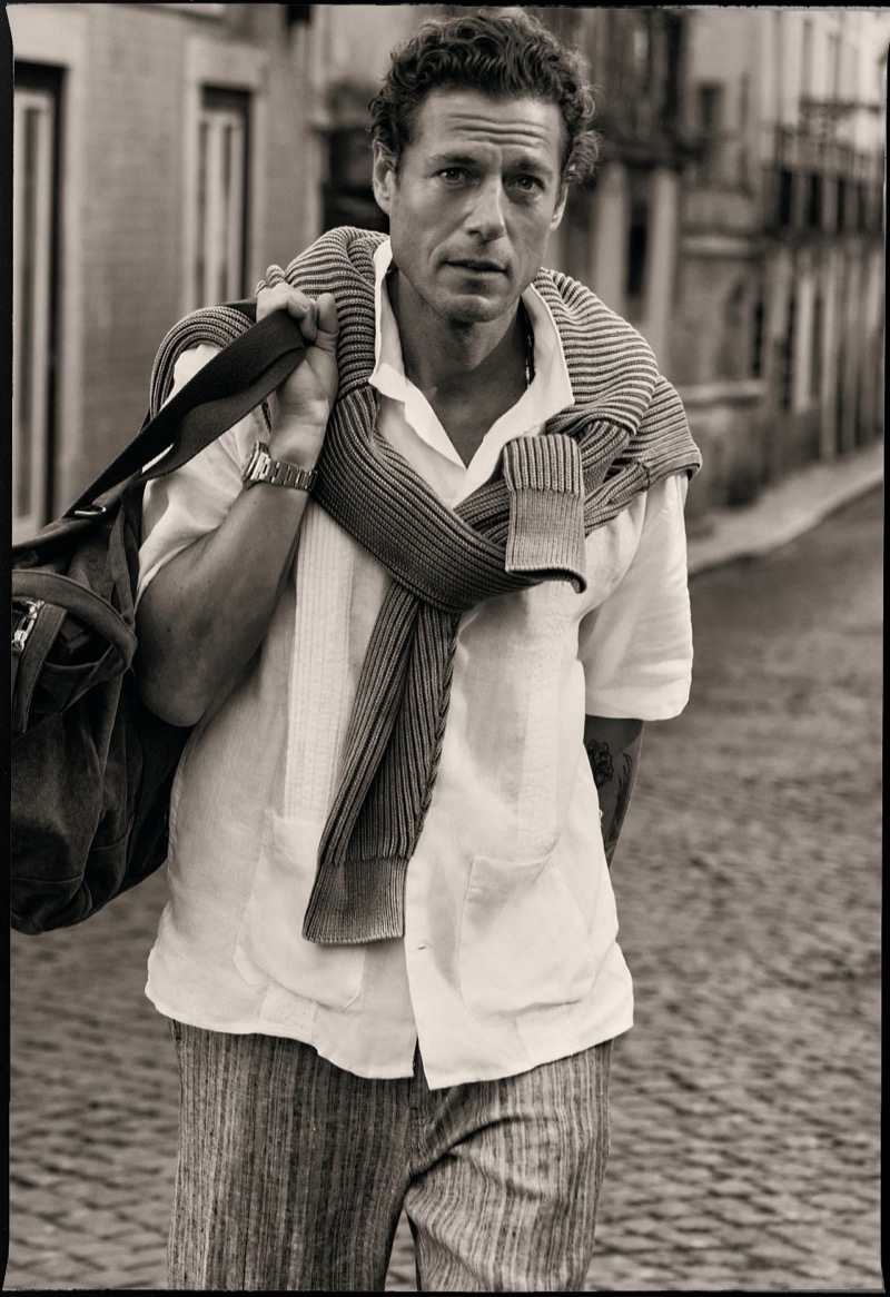 Sepia image of Jon Paul walking a cobblestone Lisbon street with a ribbed knit sweater draped over his shoulders, a white linen camp-collar shirt, striped trousers, and a canvas duffel bag on his shoulder