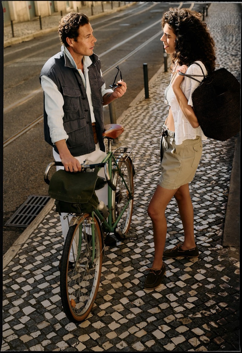 Jon Paul stands with a vintage green bicycle on a Lisbon cobblestone street talking to a female model in a slate gray quilted vest over a chambray shirt with white linen trousers and brown leather loafers