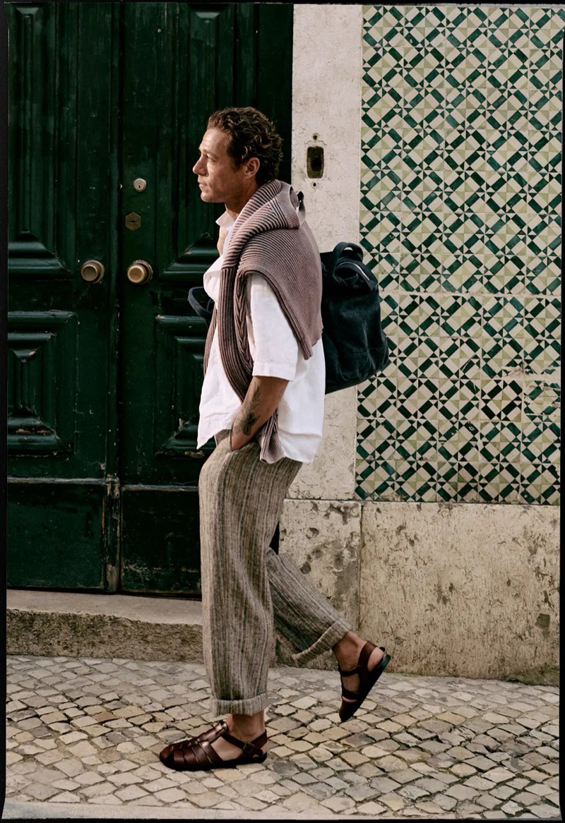 Jon Paul walks in profile past a green wooden door and green and white patterned tiles in a white linen camp-collar shirt with a knit sweater over his shoulders, striped pleated trousers, and brown leather sandals