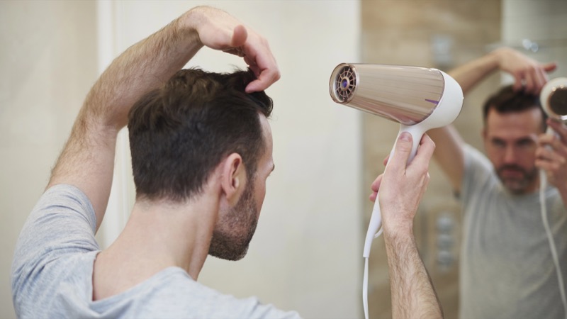 bearded man in a gray t-shirt lifts the top of his dark hair with one hand while holding a rose gold and white blow dryer at arm’s length from his head in front of a bathroom mirror