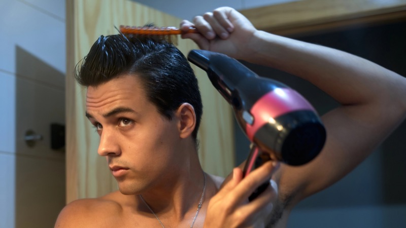shirtless man pulls a small orange comb up through the top of his dark hair while aiming a pink and black blow dryer at the roots from the side