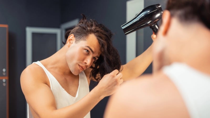 man with long dark wavy hair in a white tank top angles a black blow dryer down the length of a section of hair held in his fingers, watching himself in a dark bathroom mirror