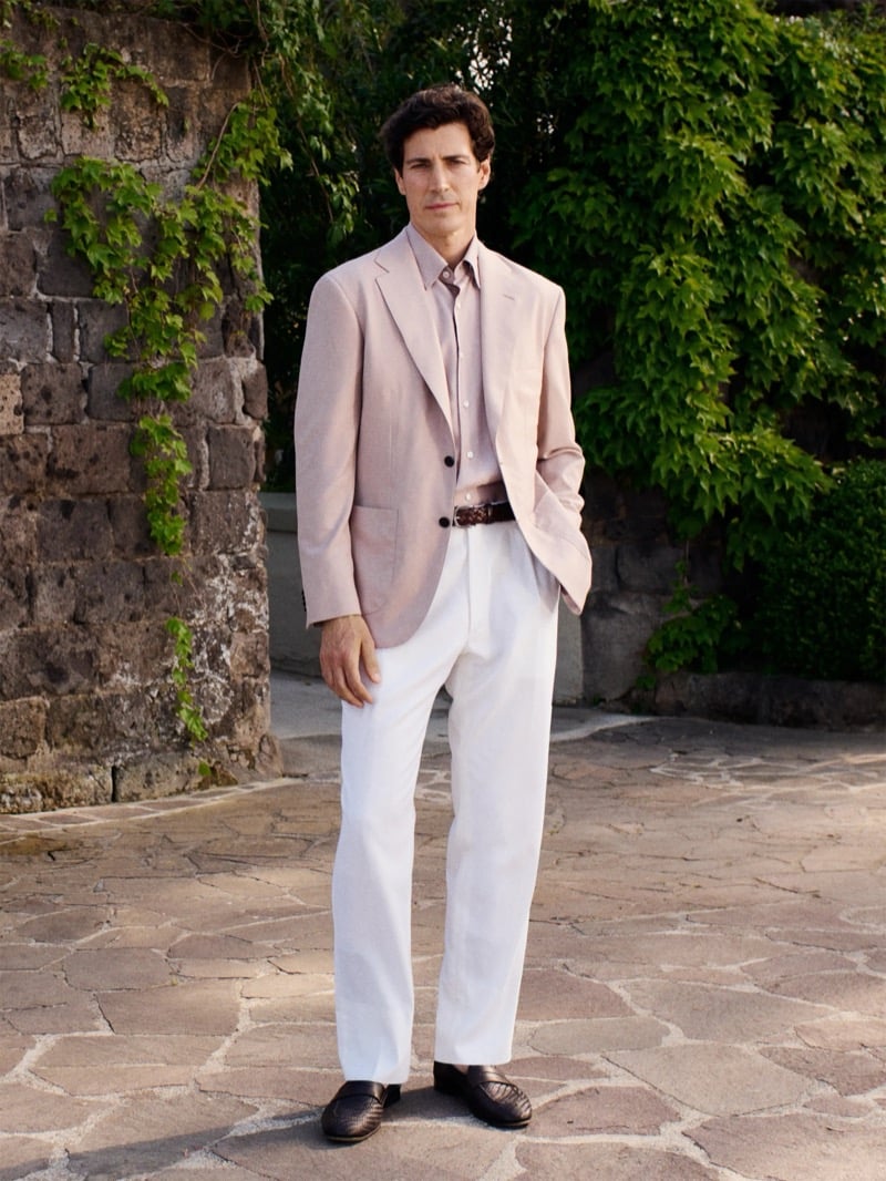 Oriol Elcacho stands on flagstone paving in a sunlit garden in front of an ivy-covered stone archway, wearing a powder pink single-breasted soft-shouldered blazer over a matching pale pink shirt and white pleated trousers cinched with a brown woven leather belt, dark woven leather loafers on his feet, dark curly hair tousled in the warm afternoon light