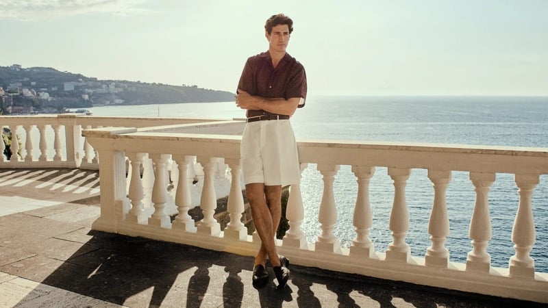 Oriol Elcacho stands with arms crossed on a sun-bleached marble terrace lined with a carved baluster railing above the Bay of Naples, the distant Sorrento coastline behind him, wearing a deep burgundy short-sleeve camp-collar shirt and pale pleated shorts cinched with a brown leather belt, dark loafers casting long shadows across the warm stone