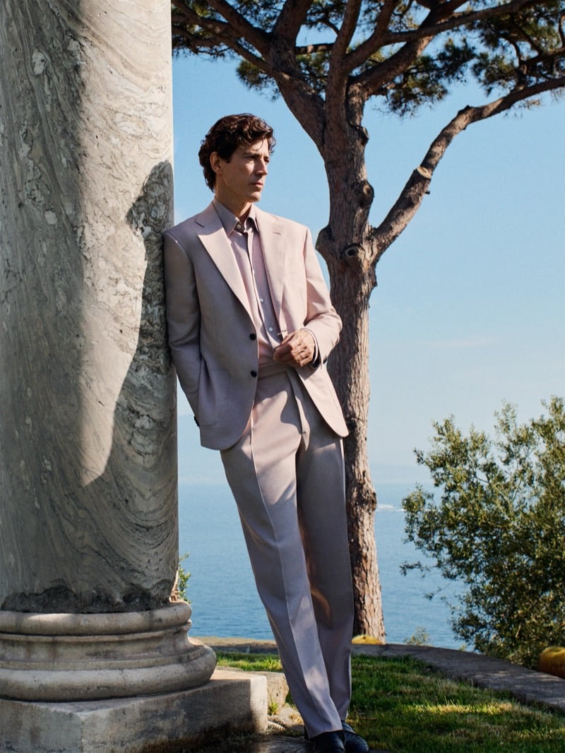 Oriol Elcacho leans against a weathered marble column under a tall Mediterranean pine on a coastal terrace with the sea stretching wide behind him, wearing a dusty powder pink soft-shouldered two-piece suit with matching pleated trousers over a pale pink dress shirt and dark leather loafers, dark curly hair and a quiet expression turned toward the camera