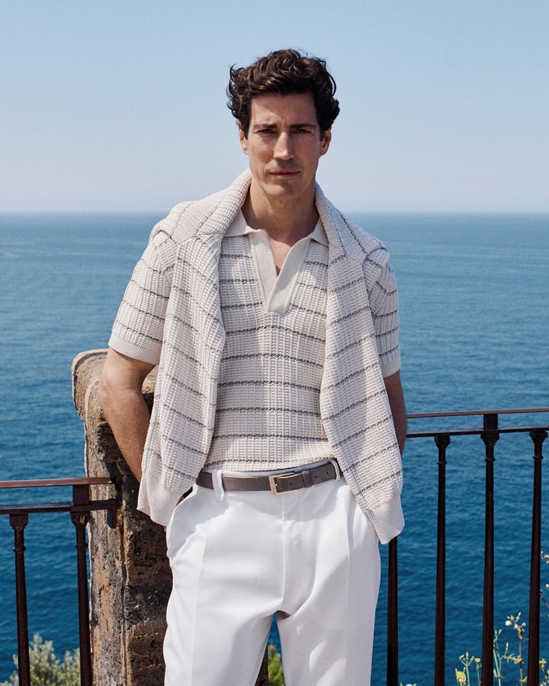 Oriol Elcacho stands at a black iron railing on a stone terrace high above the deep blue Mediterranean, one hand resting on a carved stone post, wearing a cream openwork knit polo finely striped in navy layered under a matching knit, paired with white pleated trousers and a slim brown leather belt, dark curly hair lifted in the sea breeze