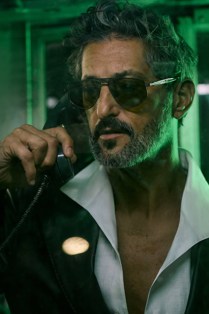 Tony angles his head toward a black corded phone receiver in slim gold-rimmed aviator sunglasses with smoke gradient lenses, a white open-collar linen shirt and a black leather jacket, washed in green sodium light