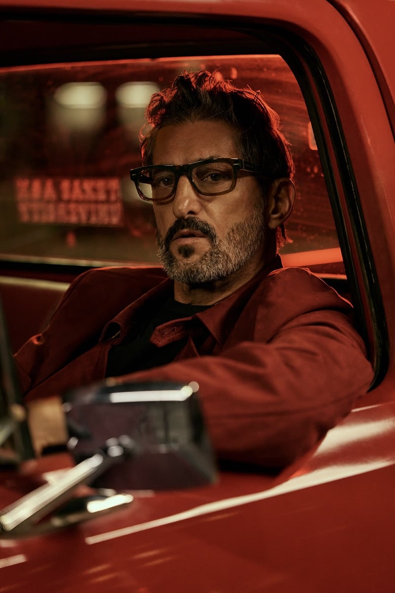 Tony sits in the cab of a red vintage pickup behind reflective glass in thick tortoise rectangular optical frames, a red corduroy shirt jacket layered over a black tee, signage glowing red in the window behind him