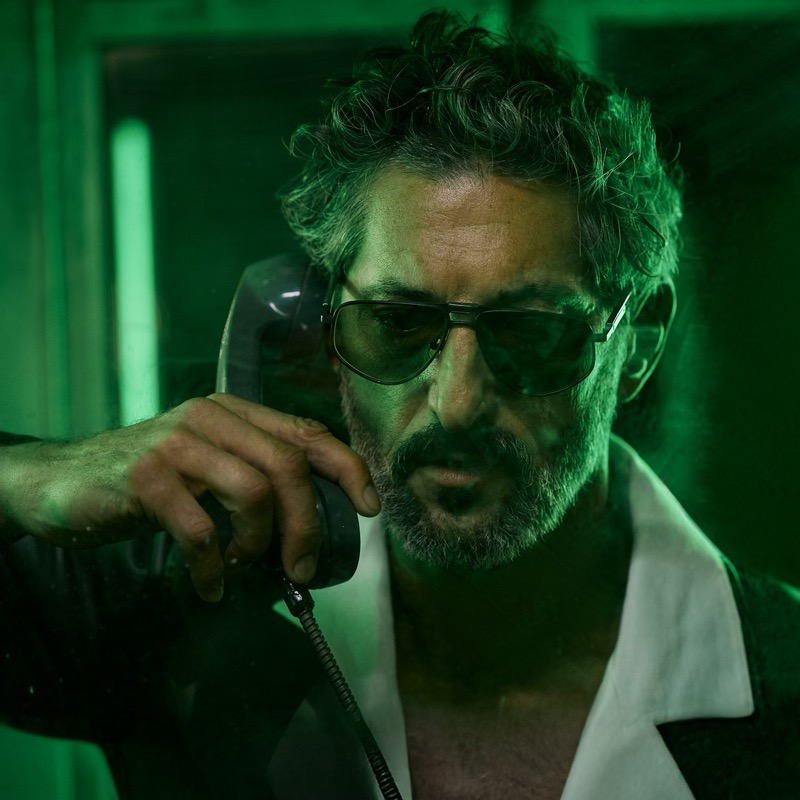 Cropped close shot of Tony in gold-rimmed aviator sunglasses lifting a black corded phone receiver to his ear, salt-and-pepper beard catching green light, white shirt collar splayed open at the chest