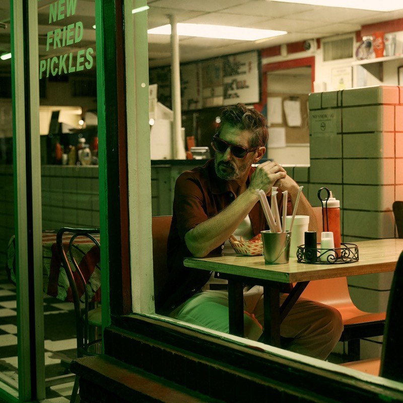 Tony seen through a diner window etched with green NEW FRIED PICKLES lettering, seated alone at an orange vinyl booth in dark sunglasses, a brown short-sleeve camp shirt and tan trousers, sipping from a paper cup with red and white straws