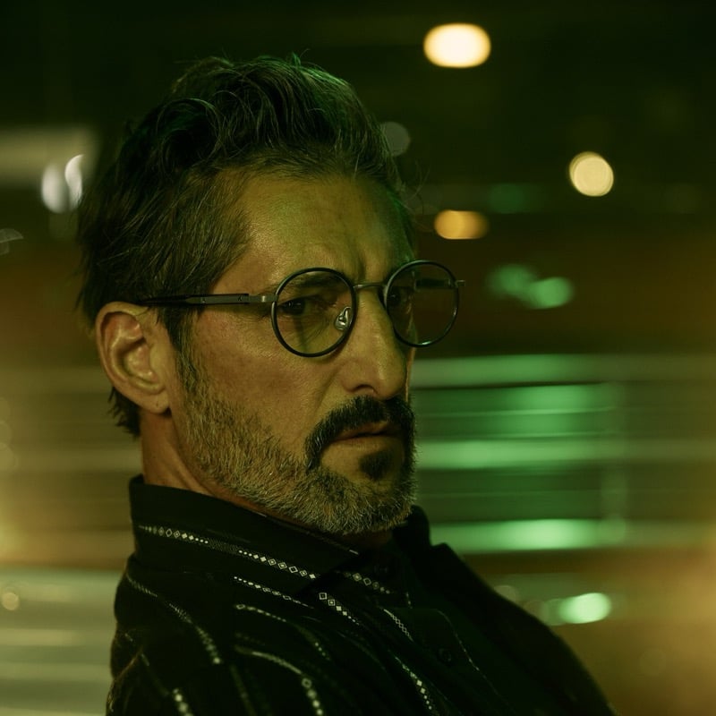 Tony turns three-quarters to camera in small round black acetate optical frames with silver hinge detail, a black geometric-print western shirt, green and amber bokeh blurring the background
