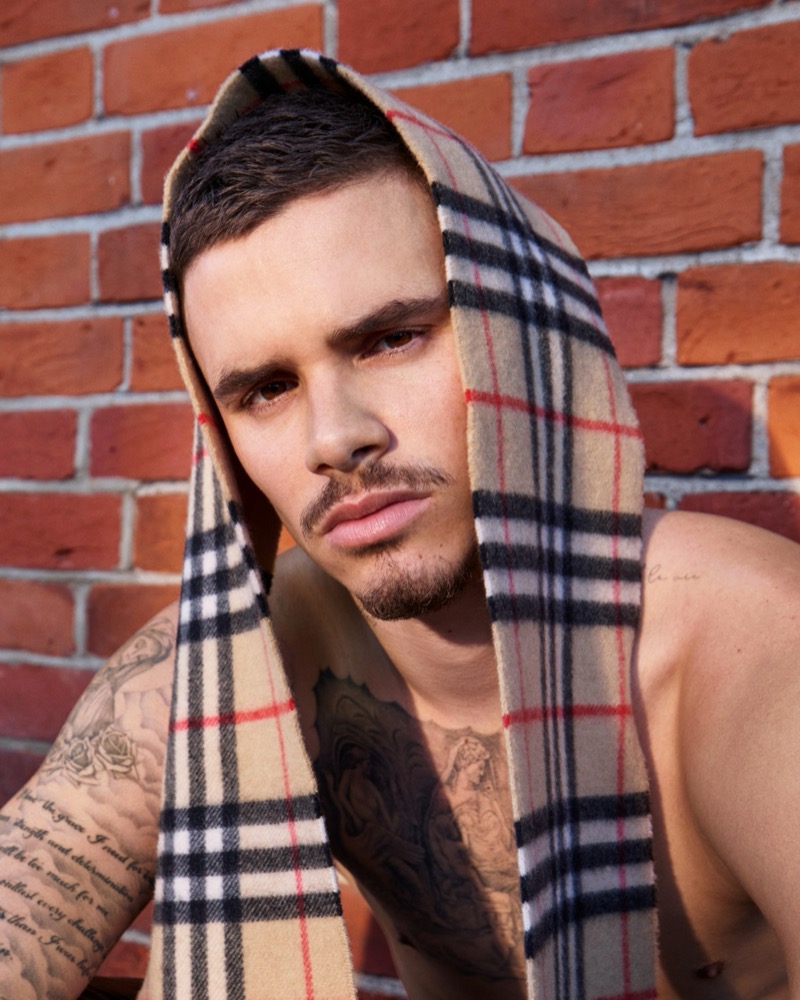 Romeo Beckham looks directly at the lens against a red brick wall with a classic Burberry check cashmere scarf draped over his head and across his bare shoulders, sleeve and chest tattoos visible