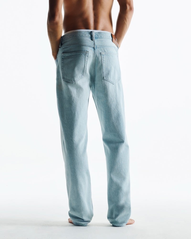 Back view studio portrait of Morgan Sahadow shirtless against a clean white backdrop in a pair of pale light wash 90s straight-leg jeans sitting low at the waist, hands tucked in the back pockets, bare feet