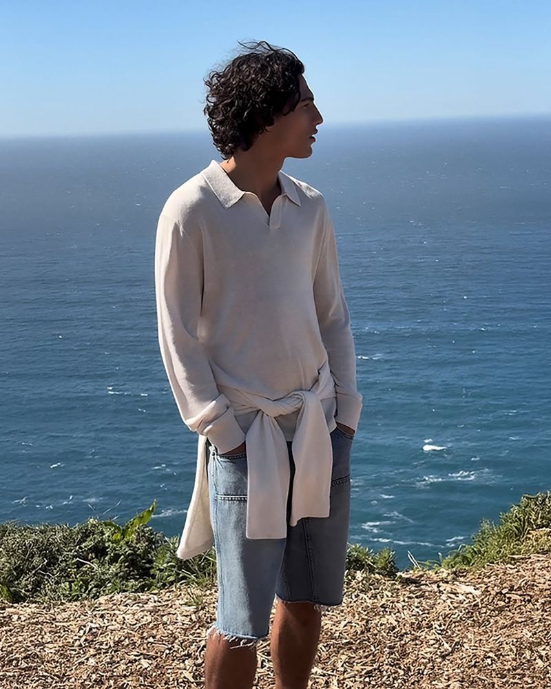 Morgan Sahadow stands on a coastal cliff above the open ocean in a relaxed oatmeal knit long-sleeve polo sweater with the spread collar open at the neck, a matching knit tied at the waist over light wash cut-off denim shorts, dark curly hair lifted by the wind