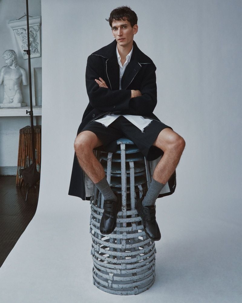 Thibaud Charon sits cross-armed atop a metal cage stool in a Camiel Fortgens oversized black wool coat with contrast white topstitched peak lapels, worn over a raw-hemmed white shirt, black shorts, gray crew socks, and heavy black leather derbies, classical plaster busts on shelves behind him