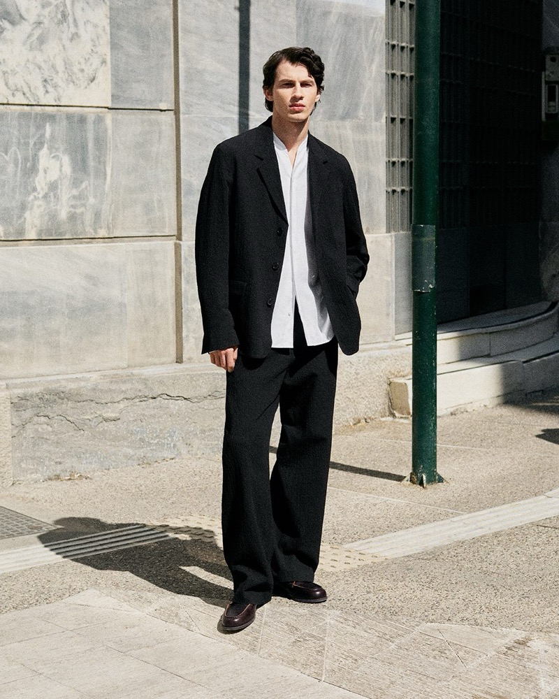 Kaplan stands on a sunlit street in a black oversized blazer, loose white shirt, dark wide-leg trousers, and brown loafers against a marble facade for the COS in the City summer 2026 edit