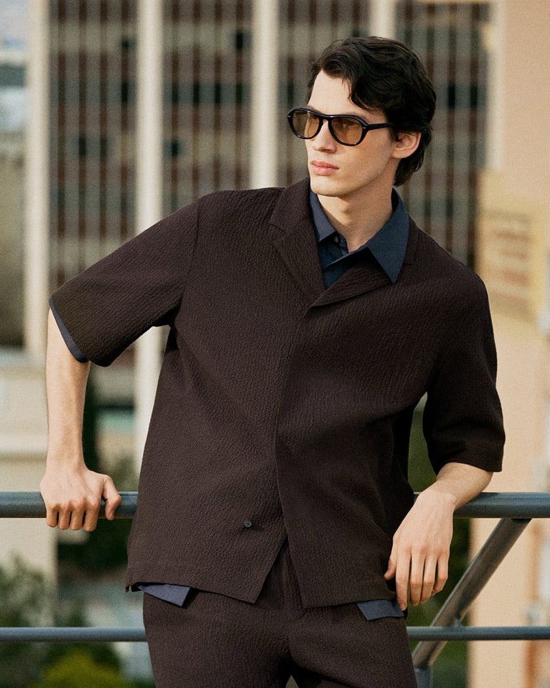 Kaplan leans on a metal rooftop railing in a dark chocolate textured short-sleeve shirt over a navy collared shirt with oblong sunglasses for the COS in the City summer 2026 edit