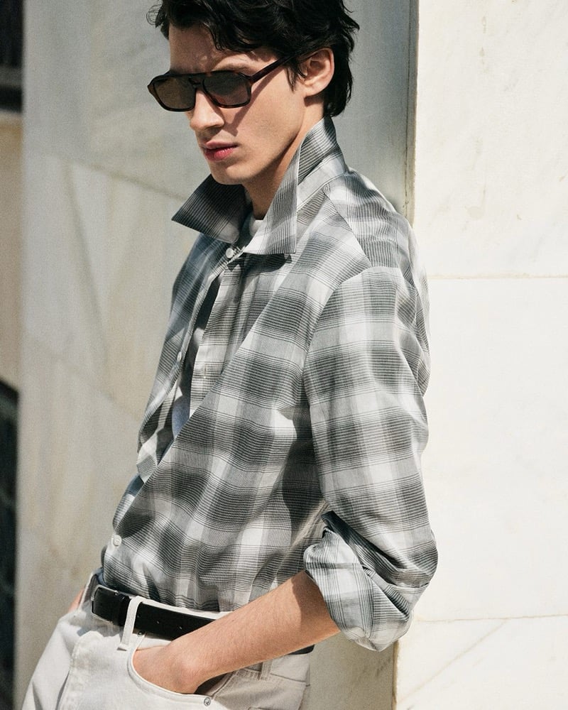 Kaplan leans against a white marble wall in a washed gray plaid long-sleeve shirt and light gray jeans with sunglasses for the COS in the City summer 2026 edit
