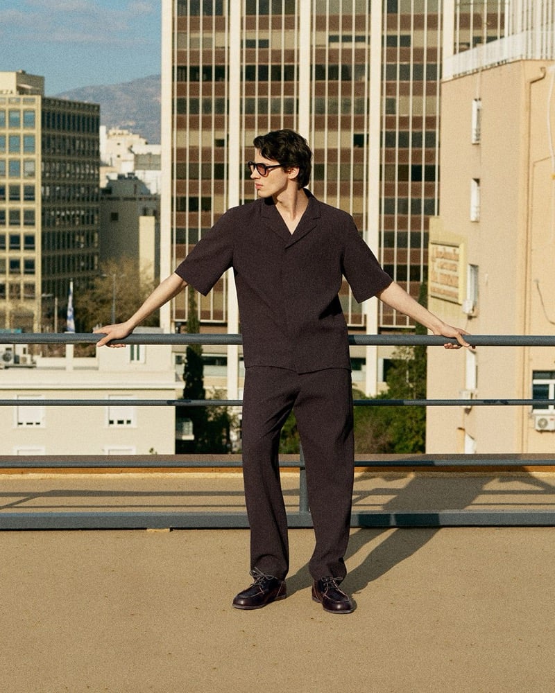 Kaplan stands on a rooftop in a dark brown textured short-sleeve camp collar shirt and matching wide-leg trousers with sunglasses and brown lace-up shoes for the COS in the City summer 2026 edit