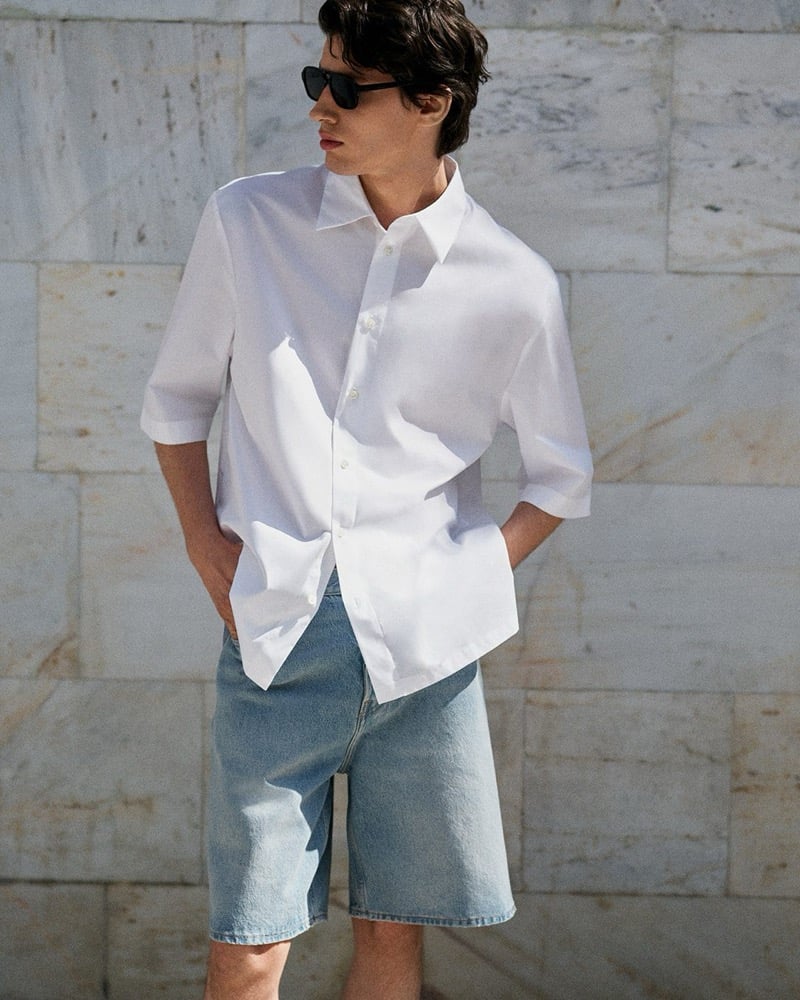 Kaplan stands against a sunlit marble wall in a white short-sleeve shirt and light wash denim shorts with black sunglasses for the COS in the City summer 2026 edit