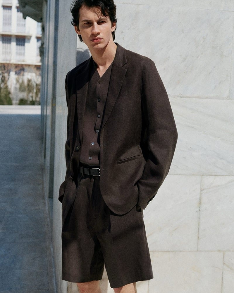 Kaplan stands against a marble wall in a dark brown linen blazer over a matching brown vest with tailored brown shorts and a black belt for the COS in the City summer 2026 edit