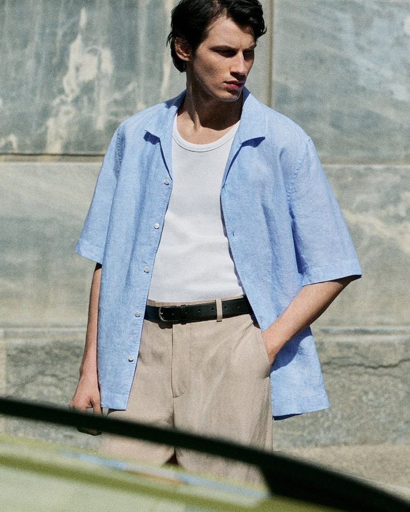 Kaplan stands on a sunlit street in a blue linen short-sleeve shirt over a white ribbed tank with beige trousers and a black belt for the COS in the City summer 2026 edit