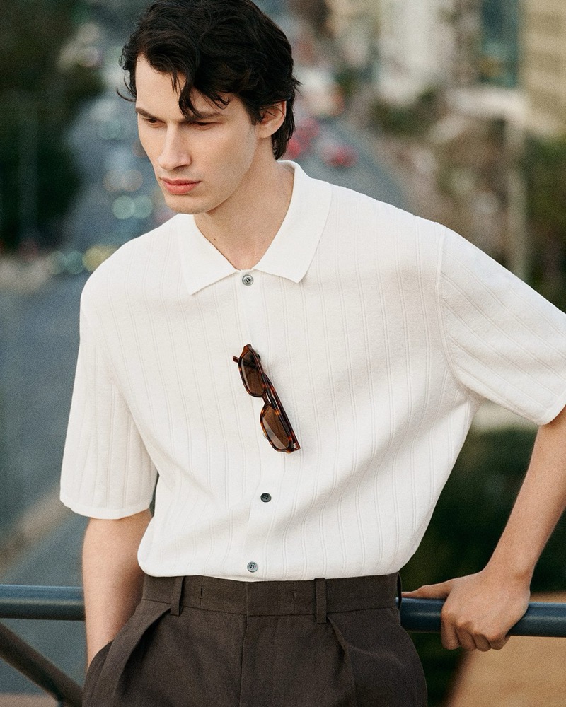 Kaplan leans on a railing above a city street in an ivory ribbed short-sleeve knit polo with tortoiseshell sunglasses tucked at the placket and dark brown pleated trousers for the COS in the City summer 2026 edit