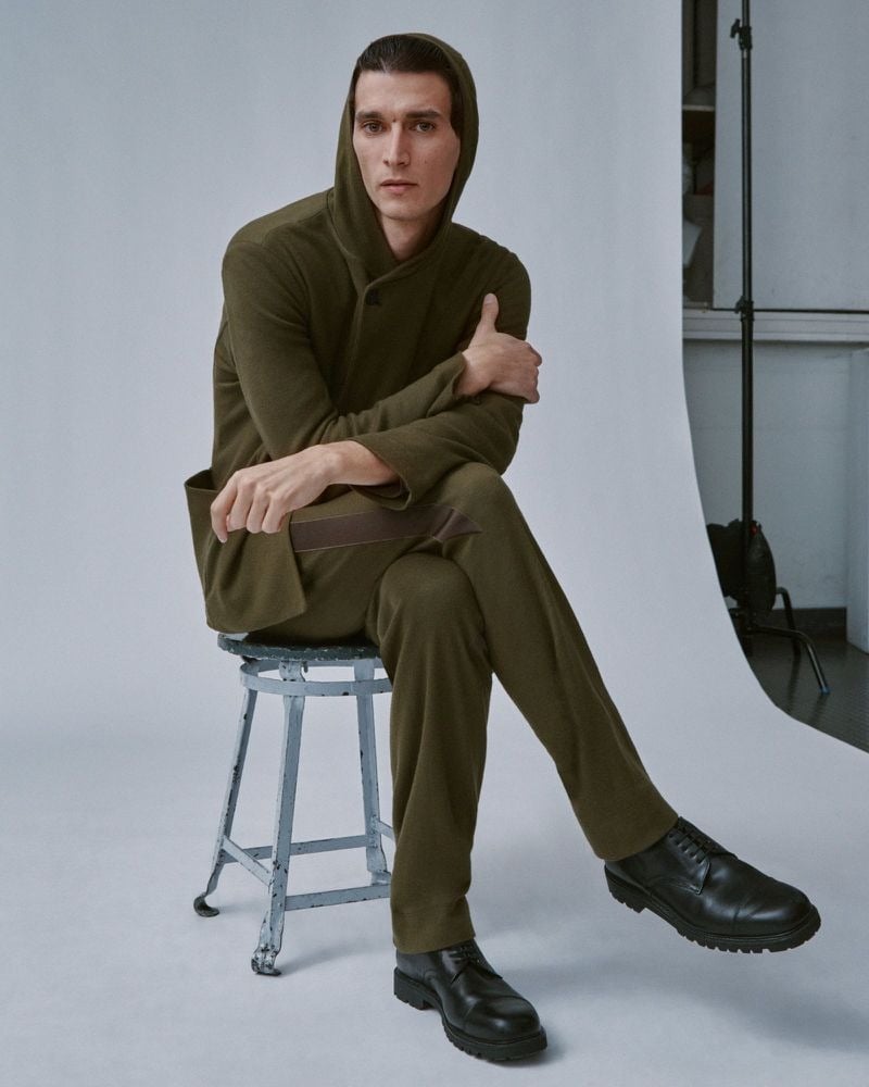 Thibaud Charon sits on a worn metal stool in a Derrick olive hooded jacket and matching olive trousers, paired with chunky black leather derbies, the hood pulled up, studio lighting visible behind