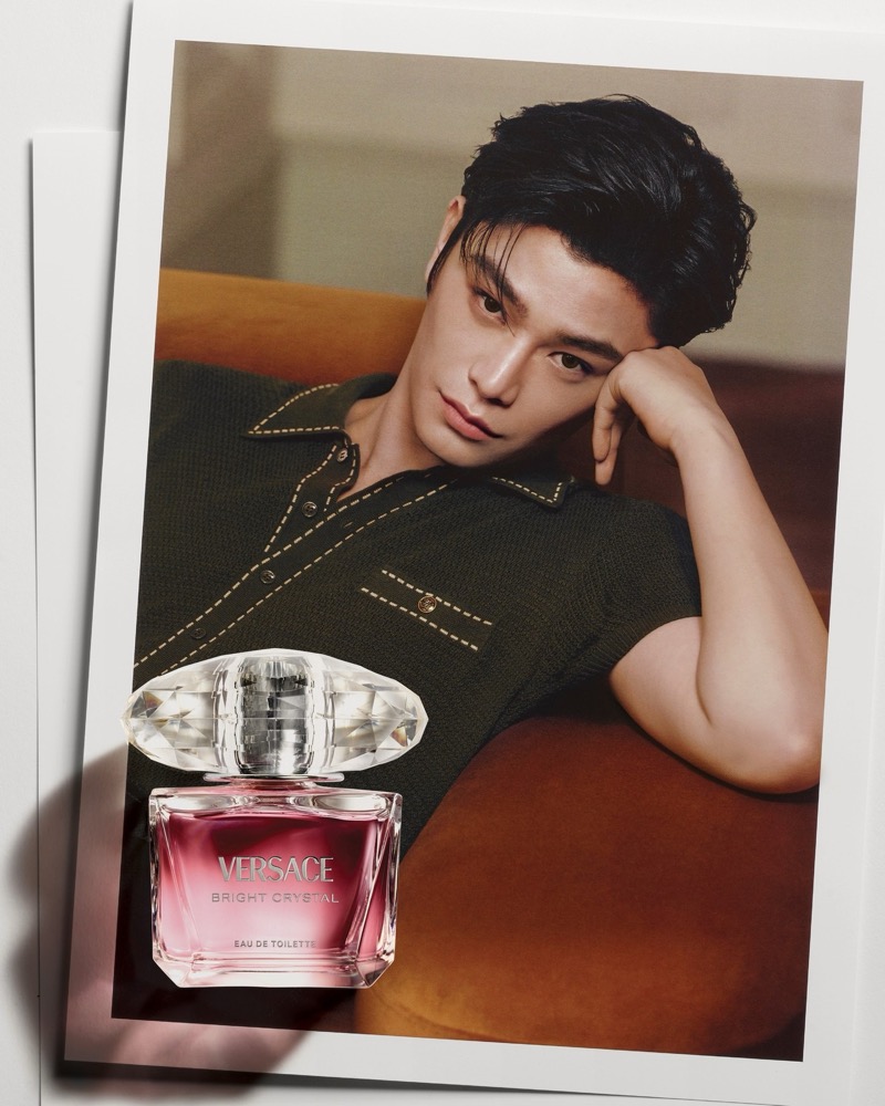 Ding Yuxi reclines across a burnt-orange velvet sofa with his chin resting in his hand, wearing a dark olive-green stitched knit polo with Medusa-engraved buttons and contrast saddle stitching, dark hair brushed back, the faceted pink Versace Bright Crystal eau de toilette flacon overlaid at the lower left of the frame