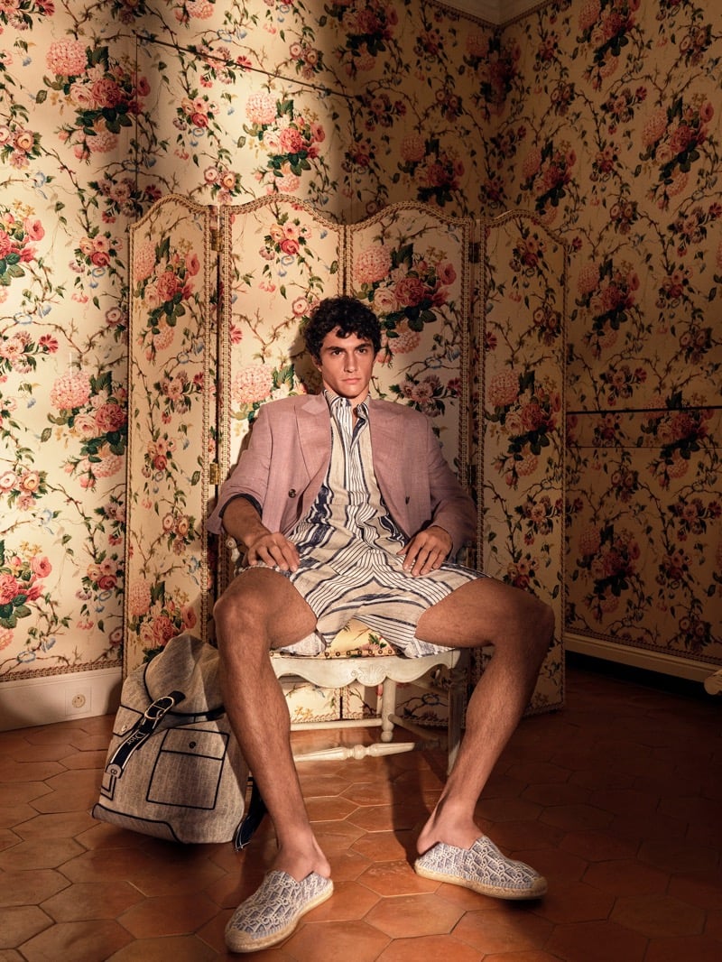 Sandro sits with legs apart on a small chair against a floral wallpaper screen in a Dior dusty pink blazer over a striped shirt and matching striped shorts with Dior Oblique espadrilles and a canvas Dior messenger bag