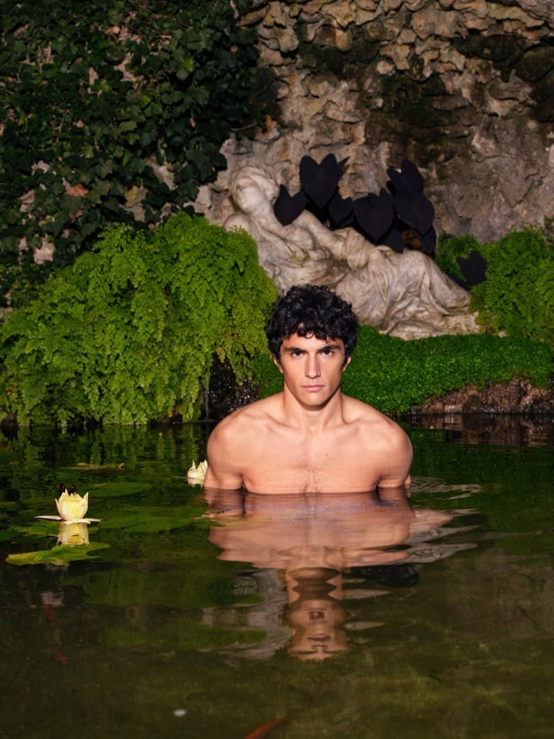 Sandro stands shirtless in a green-tinted pond surrounded by water lilies and dense ivy, with a stone statue draped in dark fabric behind him
