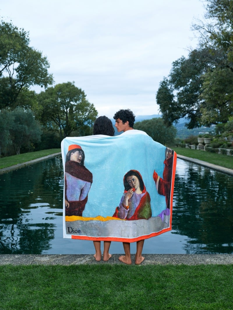 Two figures stand barefoot at the edge of a long reflecting pool wrapped together from behind in a Dior beach towel printed with a painted figurative scene and bordered in red