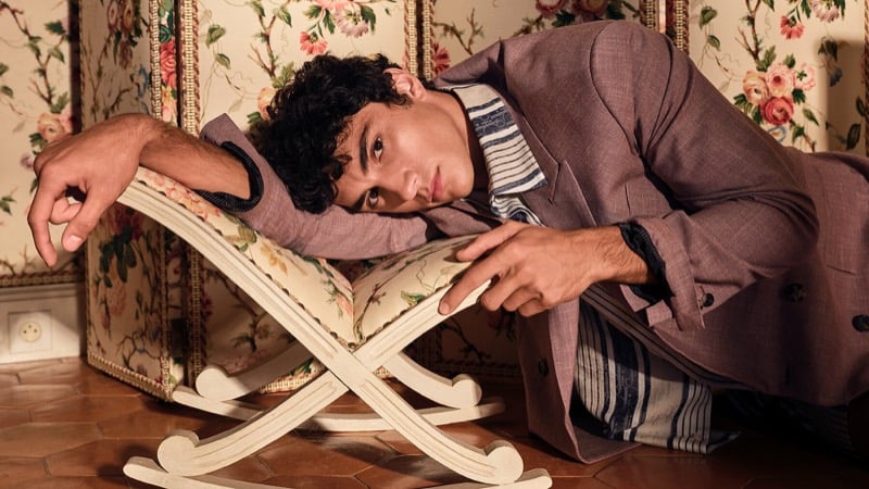 Sandro lies across a curved cream wooden bench in a Dior dusty mauve double-breasted blazer over a Breton-striped shirt against a floral wallpaper backdrop