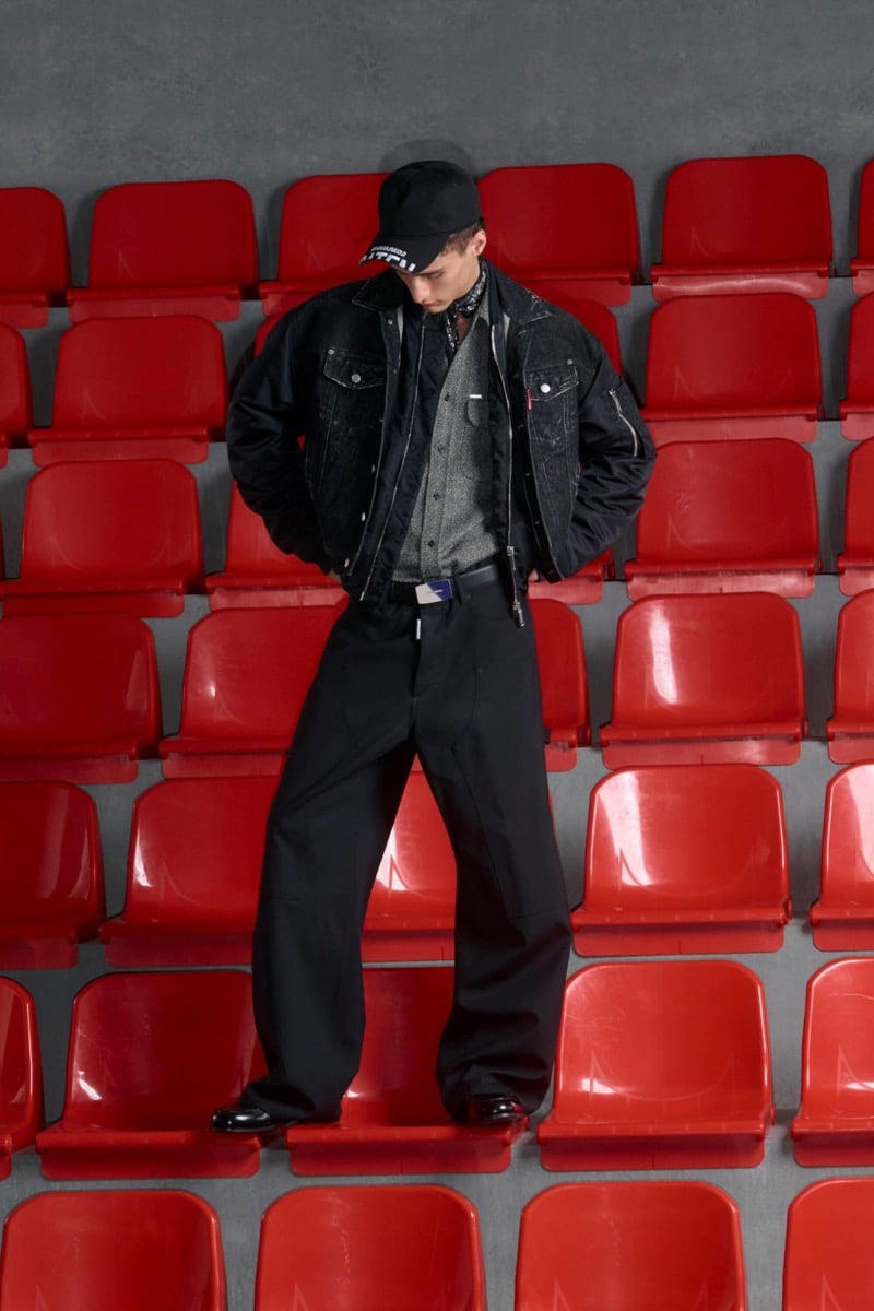 Madson Santos wears a black baseball cap and black denim trucker jacket with nylon sleeves over a gray shirt with wide-leg black wool trousers and black leather boots