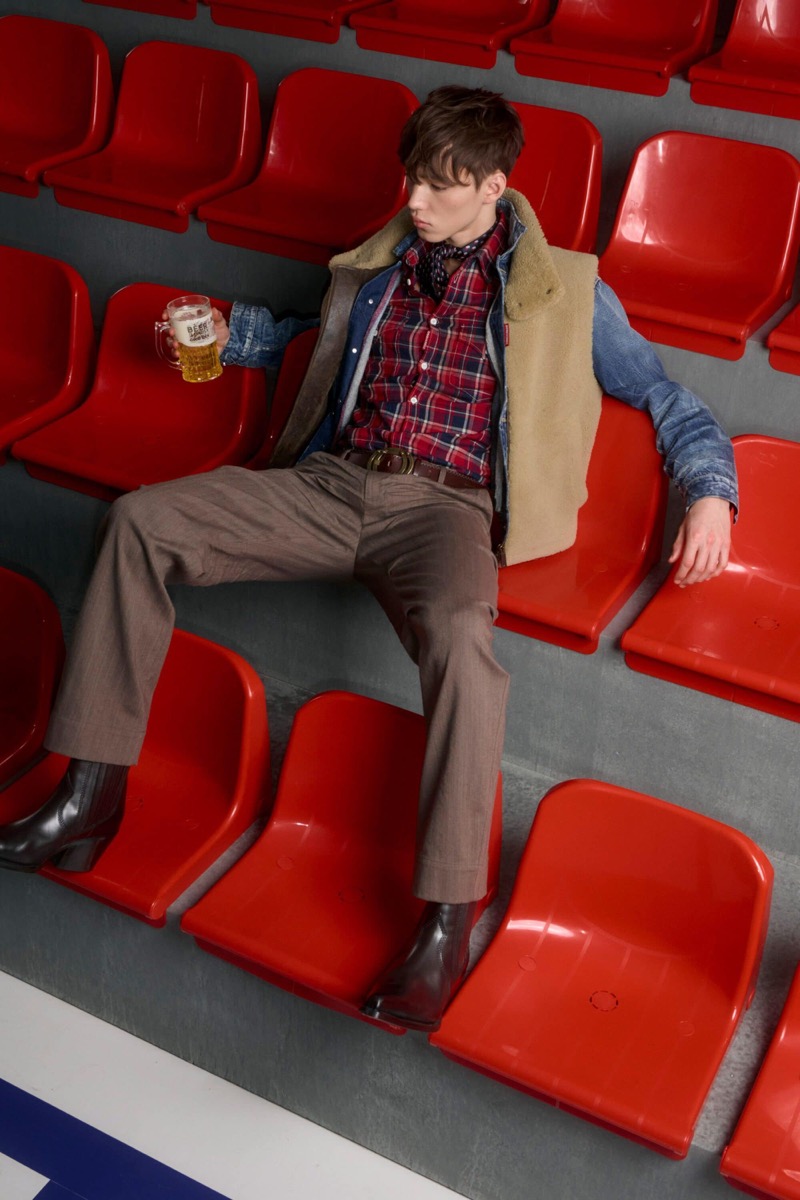 Matthew Seward wears a denim jacket with a shearling vest over a red plaid flannel shirt with a navy bandana, taupe trousers, and brown Chelsea boots, holding a beer mug