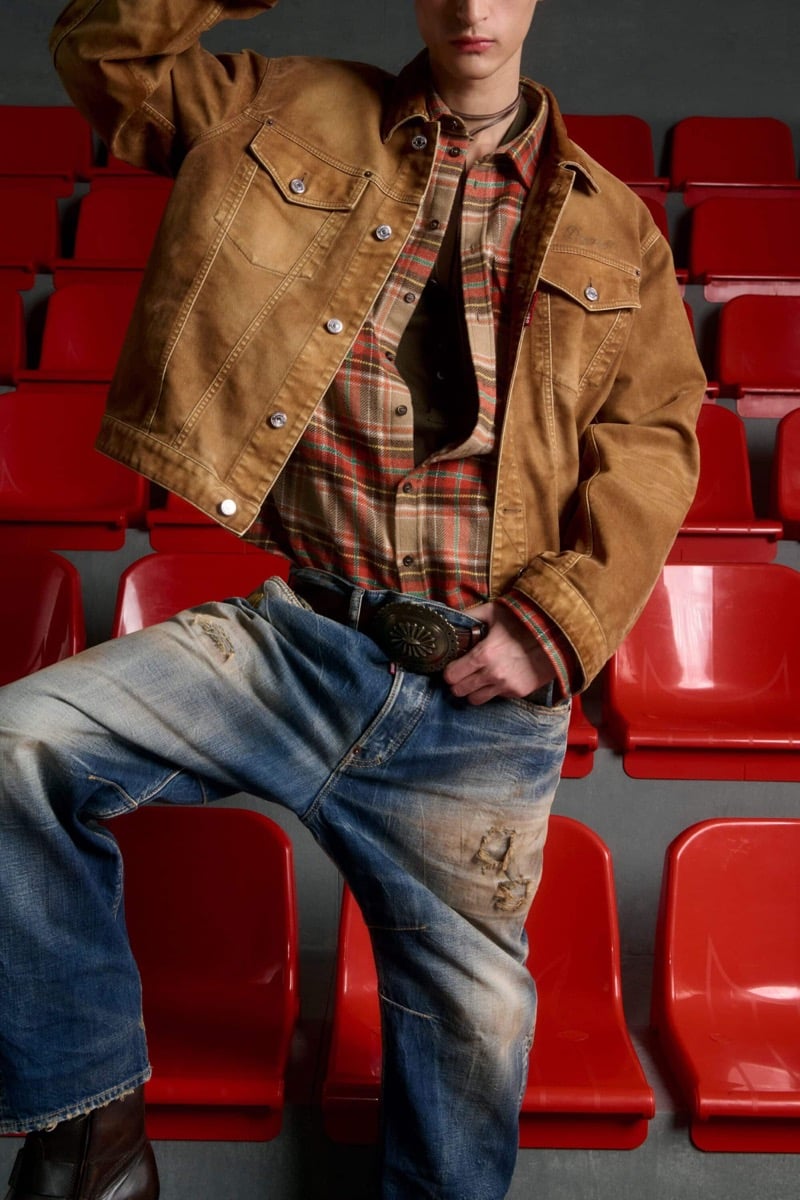 Madson Santos wears a tan canvas trucker jacket over a red and tan plaid flannel shirt with denim jeans, a tooled concho belt, and brown leather boots