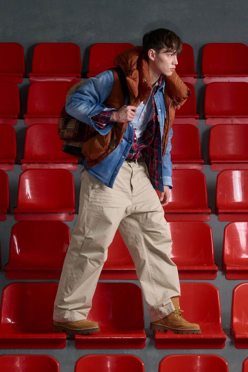 Matthew Seward wears a rust orange puffer vest over a blue denim trucker and red plaid flannel with cream chinos and tan suede work boots, carrying a plaid tartan backpack