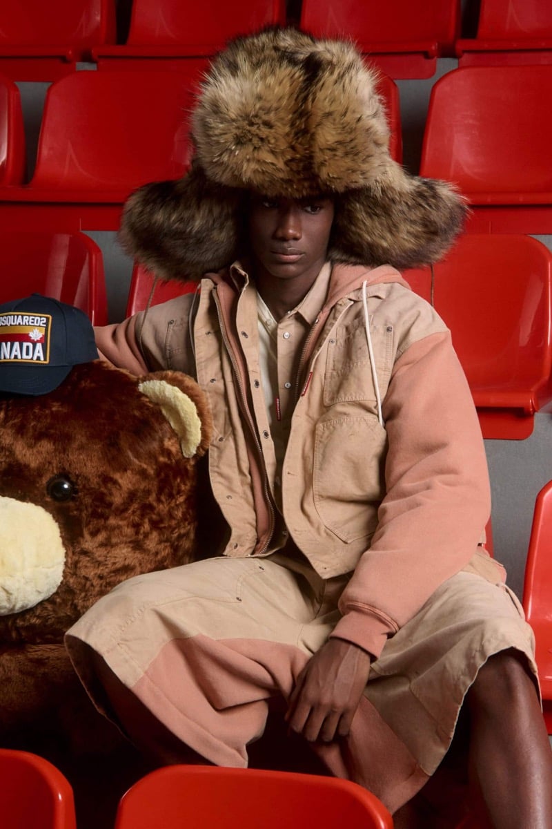 Nehemiah Kargbo wears a fur trapper hat and peach and cream colorblock canvas hooded chore jacket with matching knee-length canvas shorts, holding a navy Dsquared2 Canada cap beside a giant teddy bear