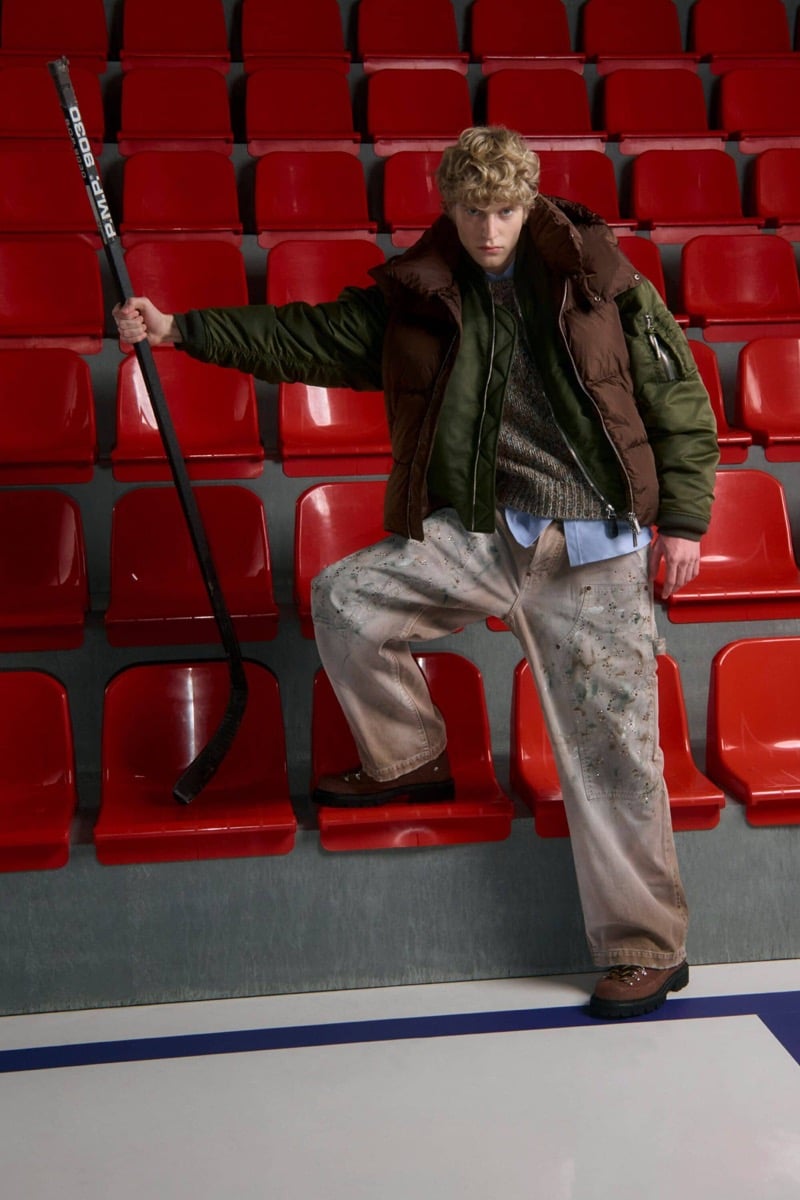 Kristaps Kallings wears a brown puffer vest over a green bomber and brown marled sweater with a blue shirt, paint-splattered baggy work pants, and brown hiking boots, holding a hockey stick