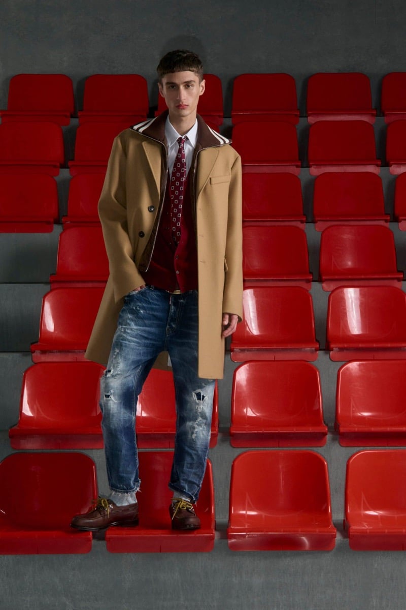 Madson Santos wears a camel double-breasted overcoat over a red cable cardigan and white shirt with a red diamond-print tie, distressed denim jeans, and brown buck shoes