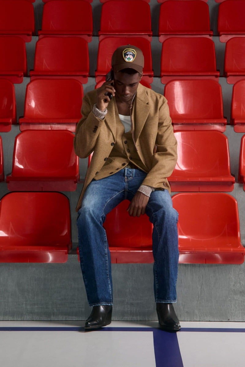 Nehemiah Kargbo wears a brown patch baseball cap and a camel wool blazer over a camel cardigan and white tank with mid-blue jeans and black Chelsea boots, holding a phone to his ear