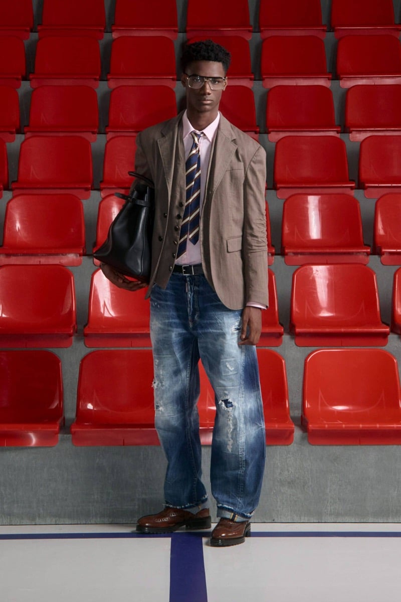 Nehemiah Kargbo wears oversized clear glasses and a taupe brown wool blazer over a waistcoat and pink shirt with a navy and gold striped tie, distressed jeans, and brown leather longwing brogues, holding a black leather bucket bag