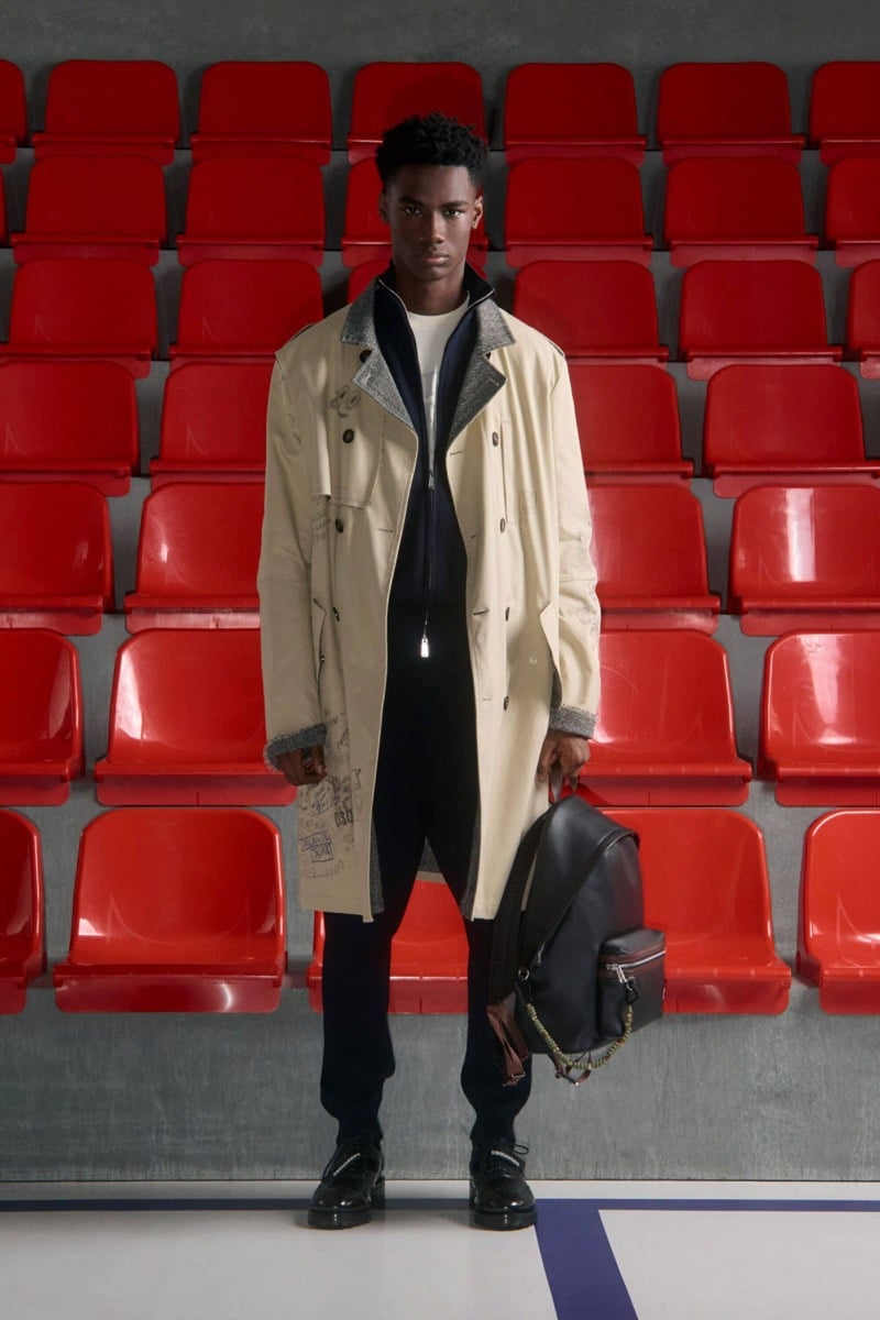 Nehemiah Kargbo wears a cream trench coat over a black zip jacket and white tee with navy track pants and black derby shoes, holding a black leather backpack with carabiner chains