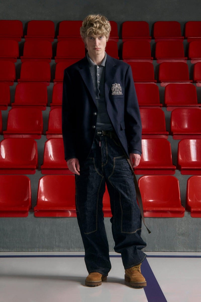 Kristaps Kallings wears a navy blazer with an embroidered crest over a ribbed polo with baggy jeans and tan suede work boots