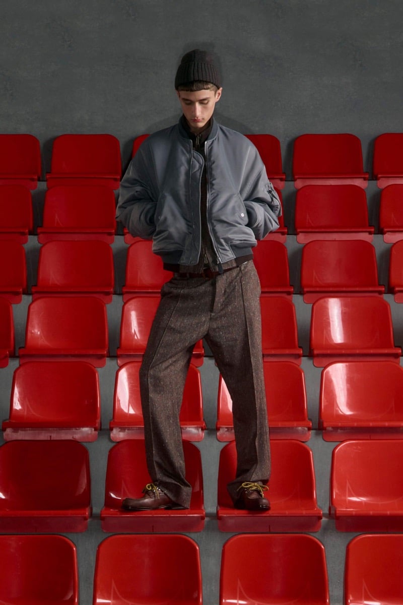 Madson Santos wears a black ribbed beanie and gray nylon bomber jacket with brown speckled tweed trousers and brown derby shoes with yellow laces