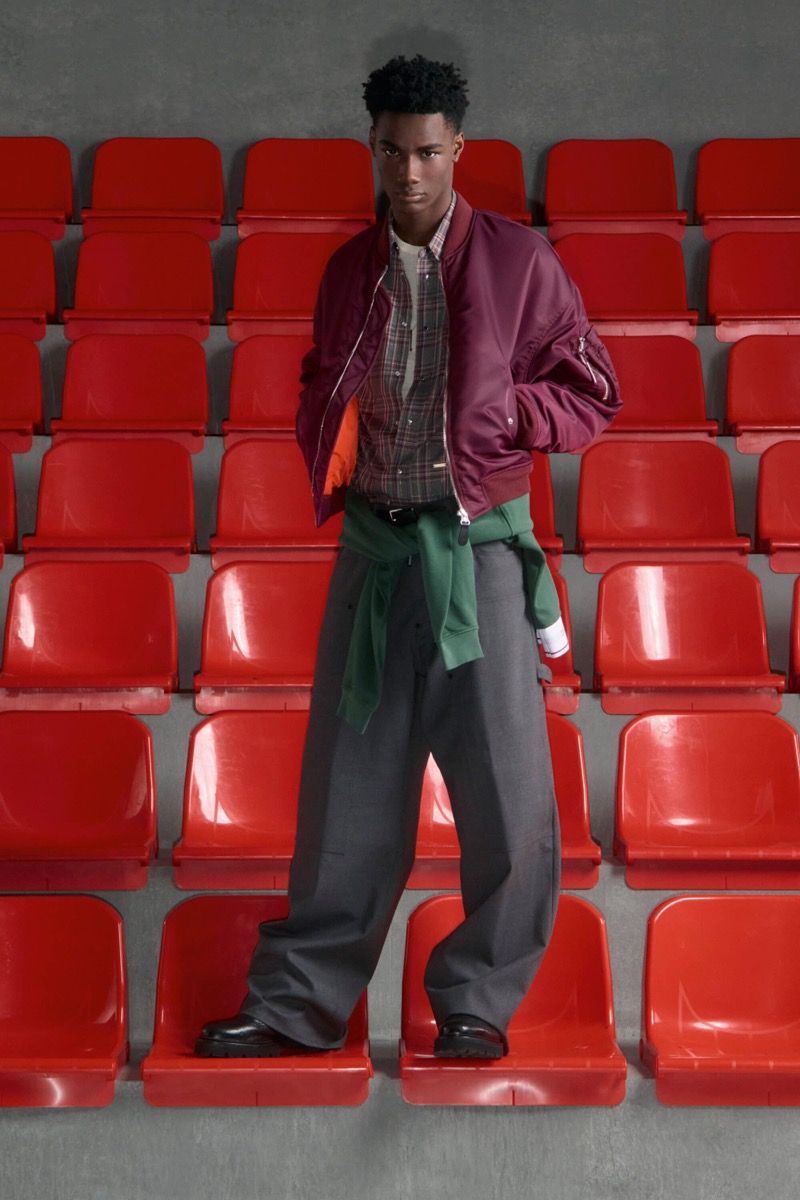 Nehemiah Kargbo wears a burgundy nylon bomber jacket with orange lining over a plaid flannel shirt and white tee with a green sweatshirt tied at the waist, gray wide-leg trousers, and black derby shoes